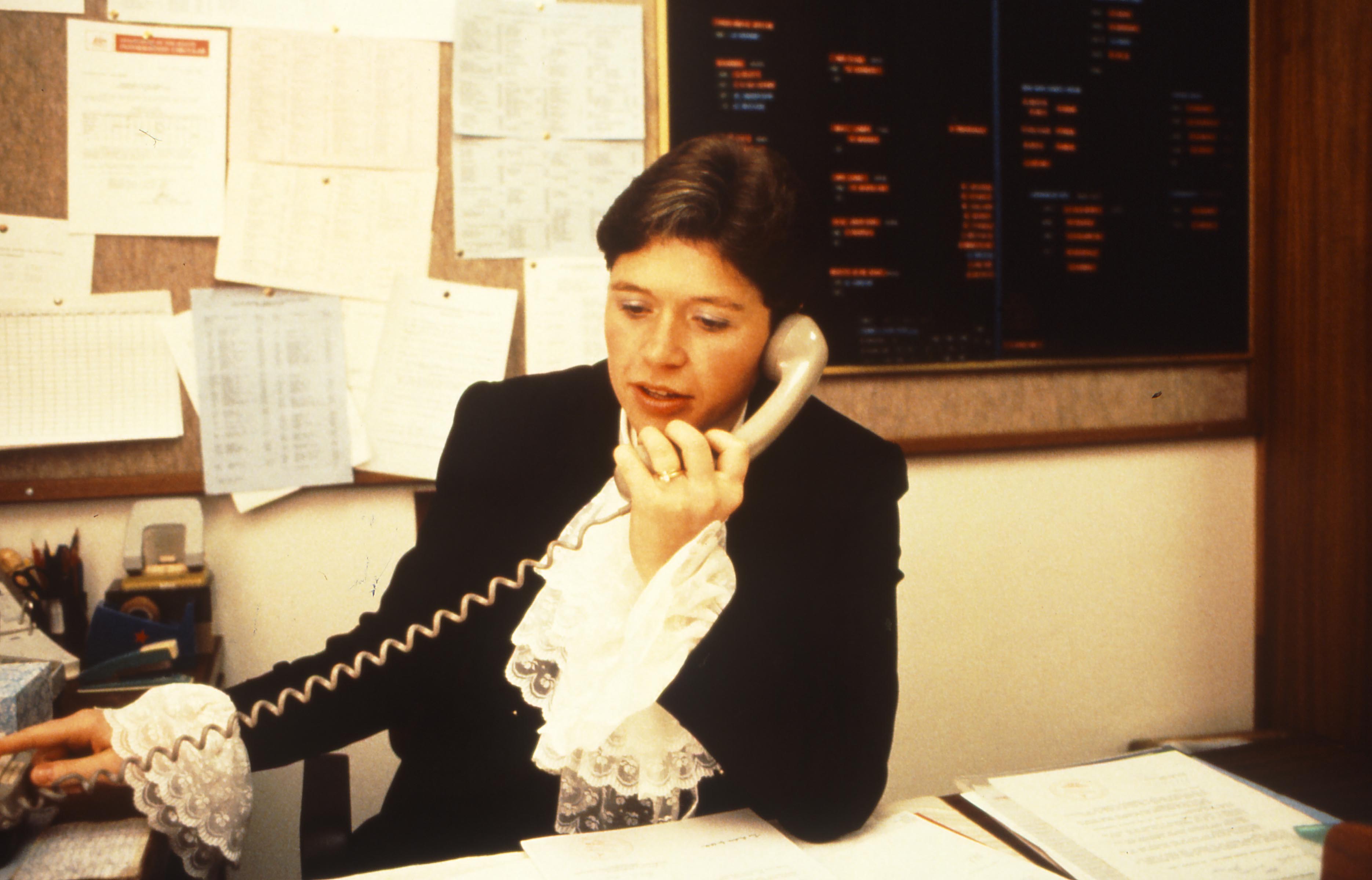 This professional colour photograph captures Andrea Griffiths making a telephone call while seated at her desk in her office in Old Parliament House. She is dressed in the ceremonial garb of the Deputy Usher of the Black Rod comprising a black suit jacket with a white lace jabot at the throat (like a frothy cravat) and white lace cuffs. There are papers on her desk and items of stationery on her desk return. The pinboard behind her is covered with notices. This professional colour photograph captures Andrea Griffiths making a telephone call while seated at her desk in her office in Old Parliament House. She is dressed in the ceremonial garb of the Deputy Usher of the Black Rod comprising a black suit jacket with a white lace jabot at the throat (like a frothy cravat) and white lace cuffs. There are papers on her desk and items of stationery on her desk return. The pinboard behind her is covered with notices.