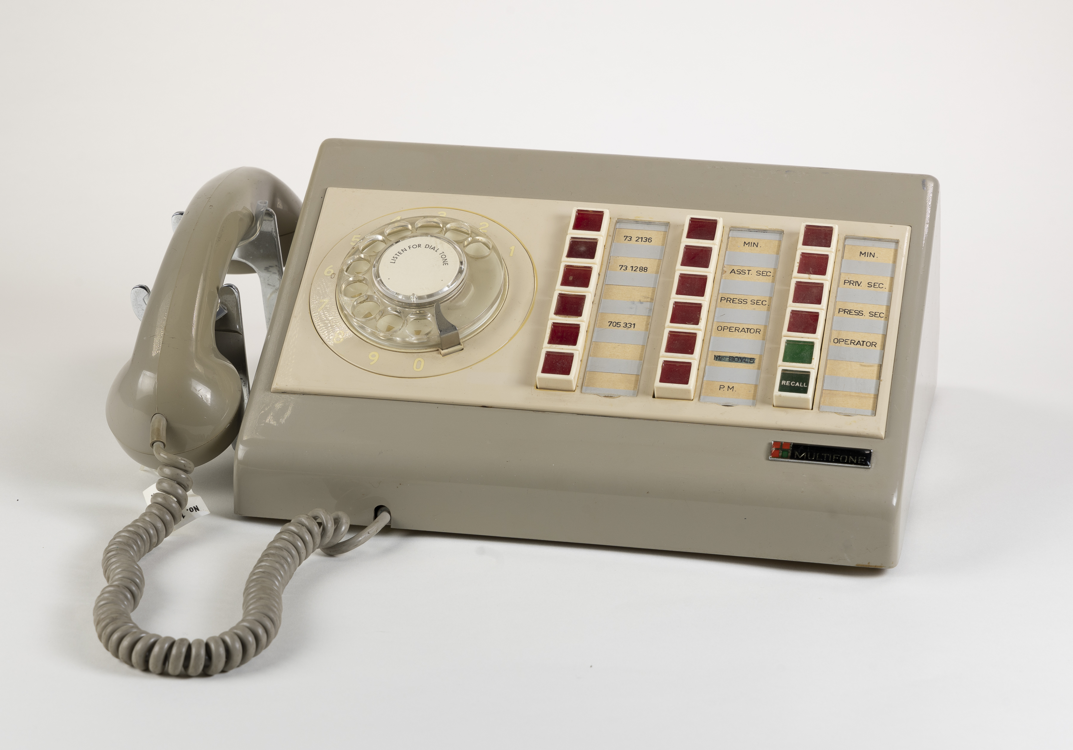 A light taupe plastic Multifone phone with 16 red buttons, 2 green buttons and a manual hand dial. The receiver sits on the side with a curly rubber cord. A light taupe plastic Multifone phone with 16 red buttons, 2 green buttons and a manual hand dial. The receiver sits on the side with a curly rubber cord.