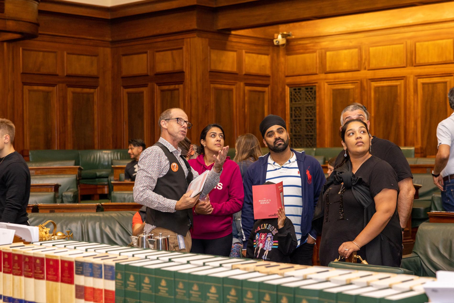 A tour guide points to something above a group of people listening to his tour. They are in the House of Representatives Chamber with green leather chairs behind them and leather bound books stacked on a central table in front of them. A tour guide points to something above a group of people listening to his tour. They are in the House of Representatives Chamber with green leather chairs behind them and leather bound books stacked on a central table in front of them.