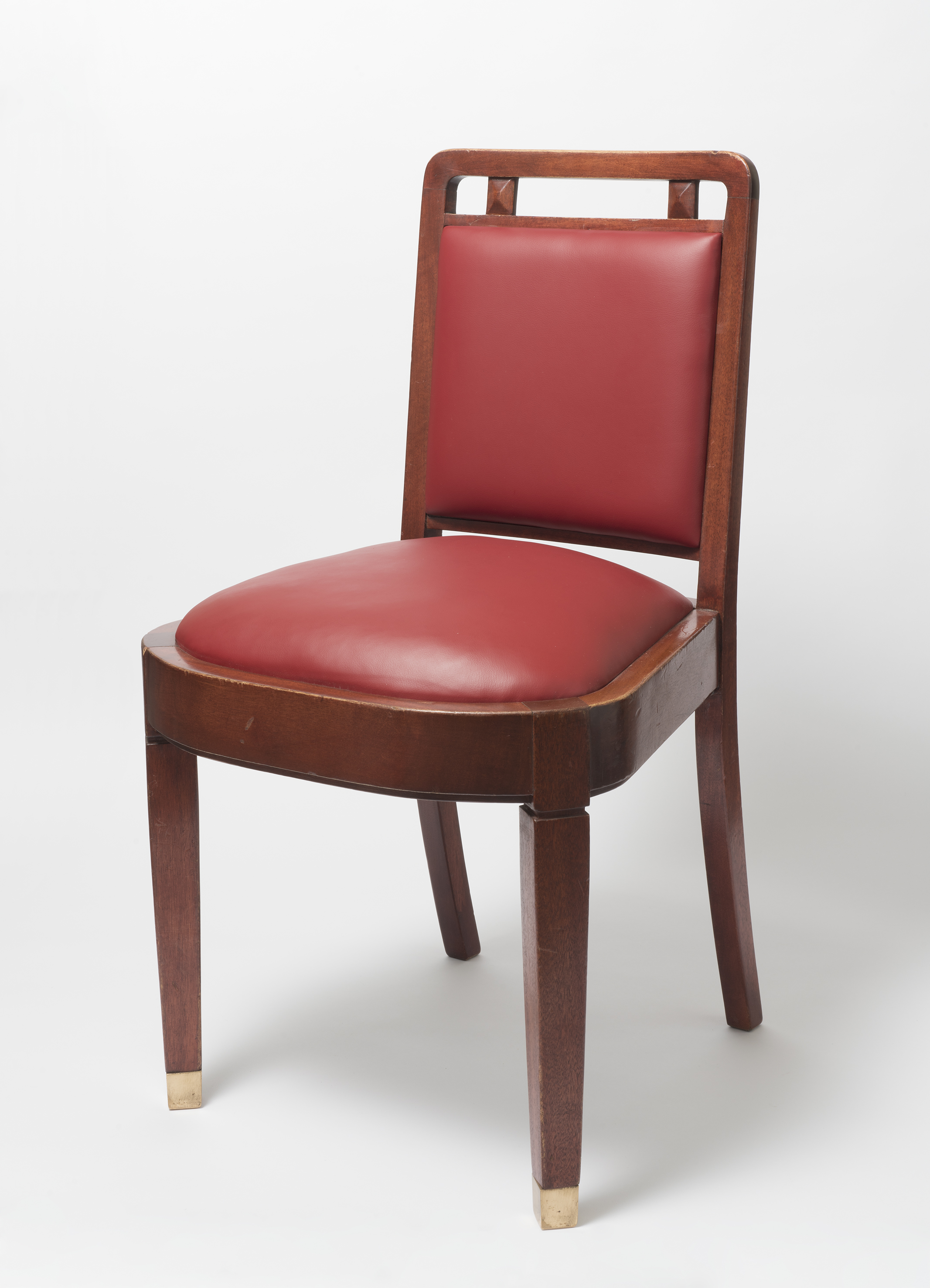 A wooden chair with cherry red leather seat cushion and back, and square legs with protective brass caps on the base of each leg. 