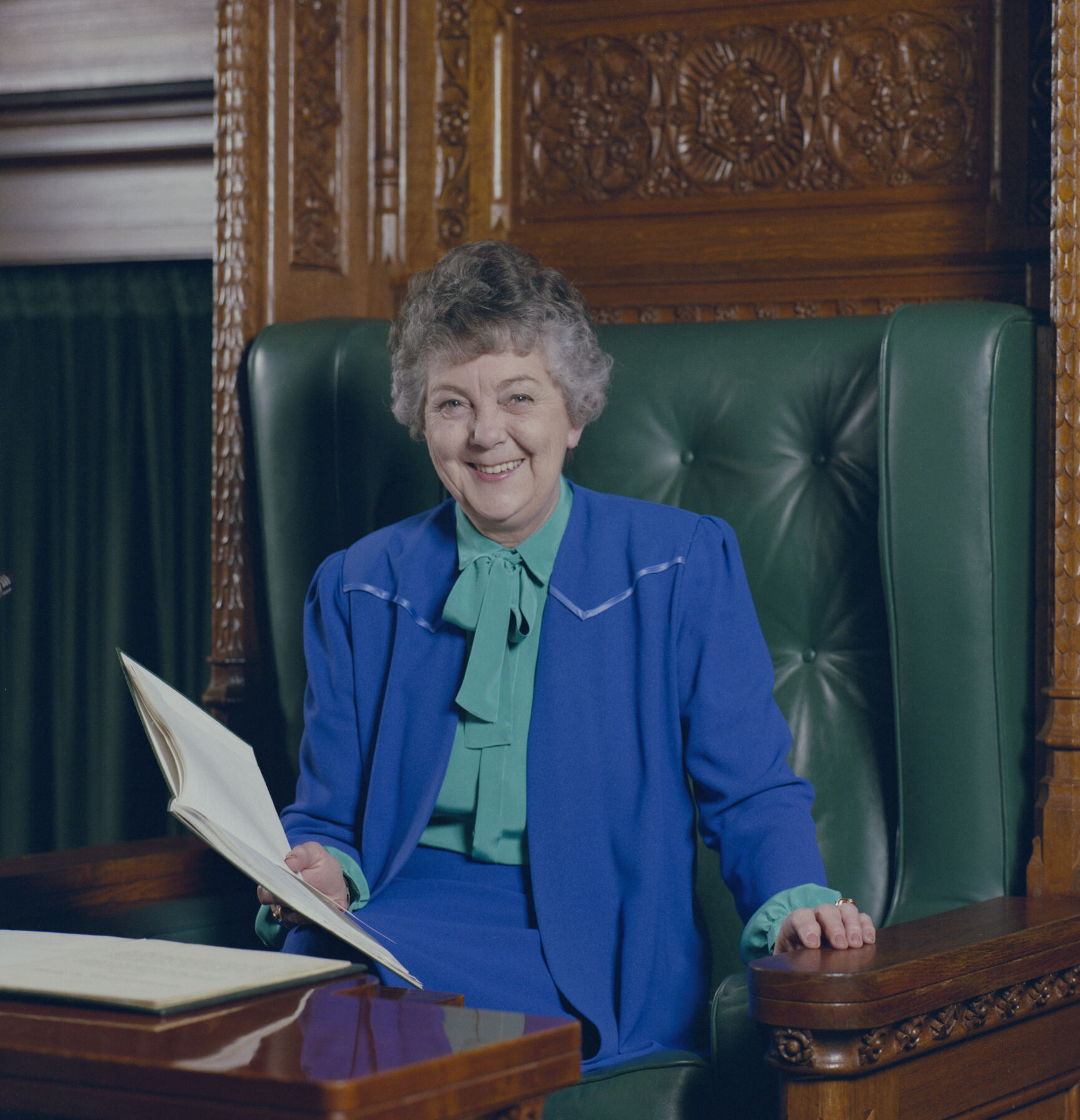A colour photograph of Joan Child wearing a blue suit sitting on the green leather and timber Speaker's Chair. A colour photograph of Joan Child wearing a blue suit sitting on the green leather and timber Speaker's Chair.