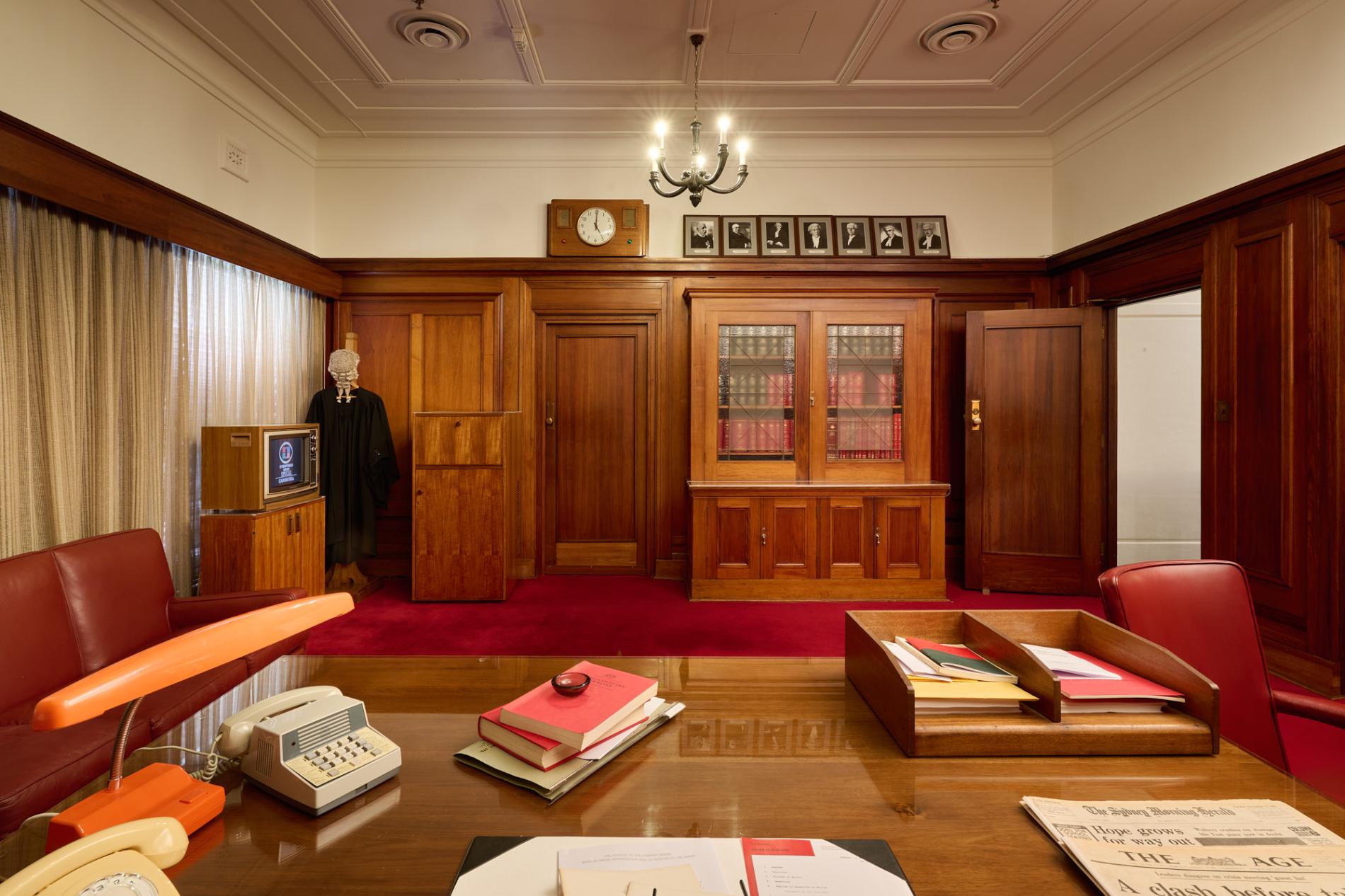 An office at Old Parliament House recreated to the 1970s with a wooden desk with an orange lamp, two phones, red-leather books and newspapers. There is red carpet, wooden bookcases and a wooden television set.