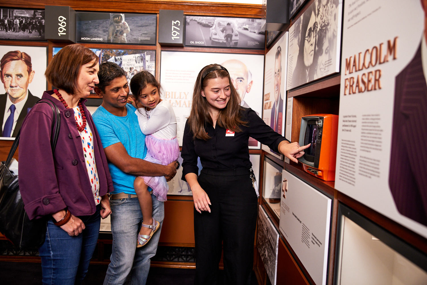 A family and a museum staff member look at an old-fashioned orange television set in an exhibition featuring Australian prime ministers at Old Parliament House.