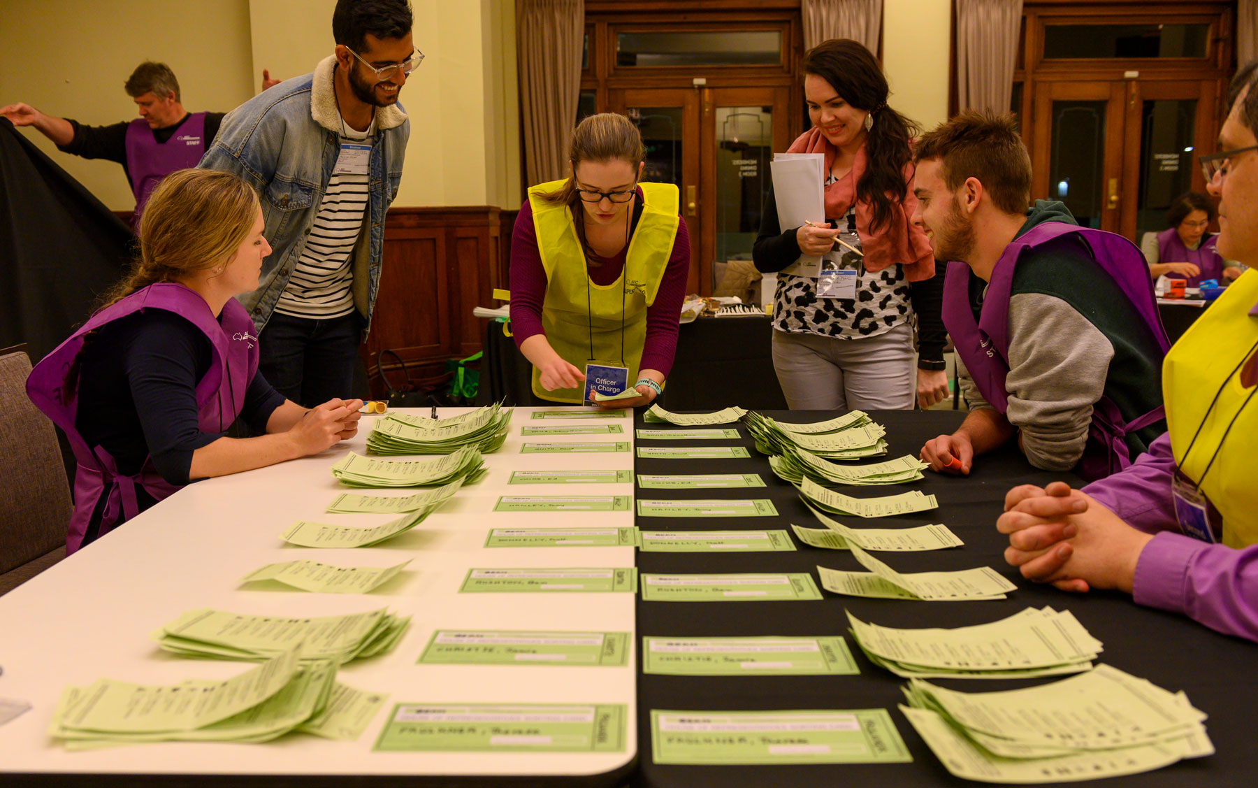 Officials in yellow and purple vests count green ballot paper votes on a table in Old Parliament House. Officials in yellow and purple vests count green ballot paper votes on a table in Old Parliament House.