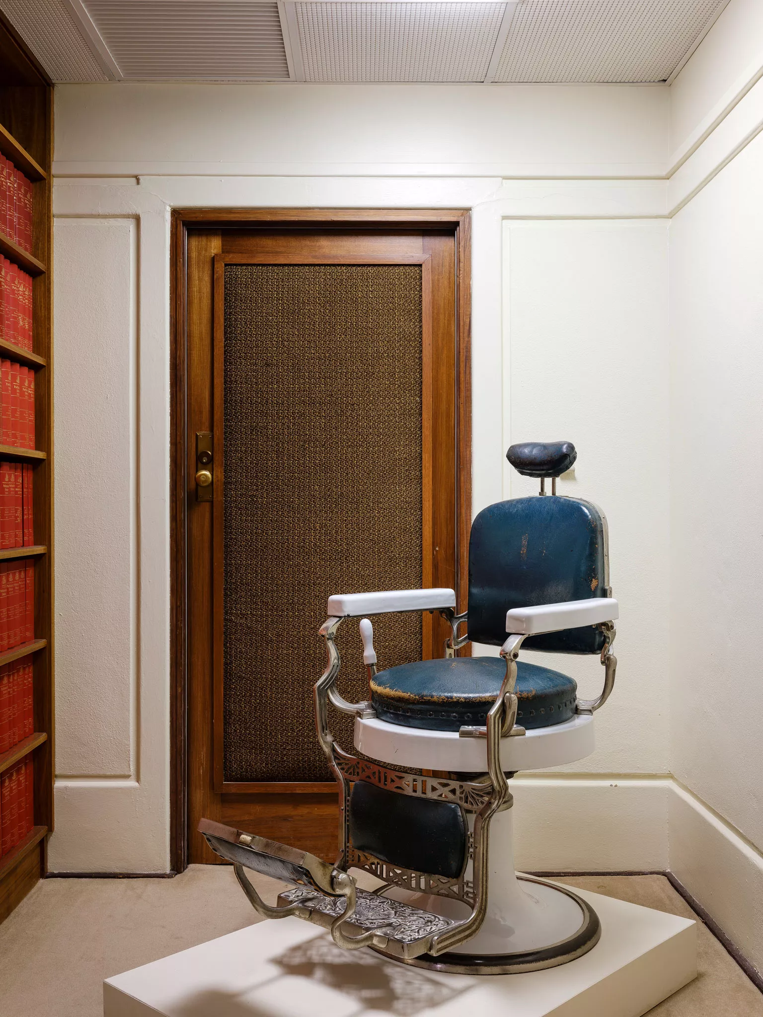 A blue leather barber's chair with white handles displayed in a room with a brown wooden door behind it. A blue leather barber's chair with white handles displayed in a room with a brown wooden door behind it.